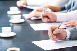 Close-up of masculine hand holding ballpoint over business document on background of human hands with cups of coffee near by